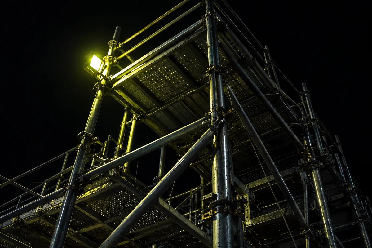 Steel scaffolding against a black sky, shot from below, with one lime work light clamped to a pole